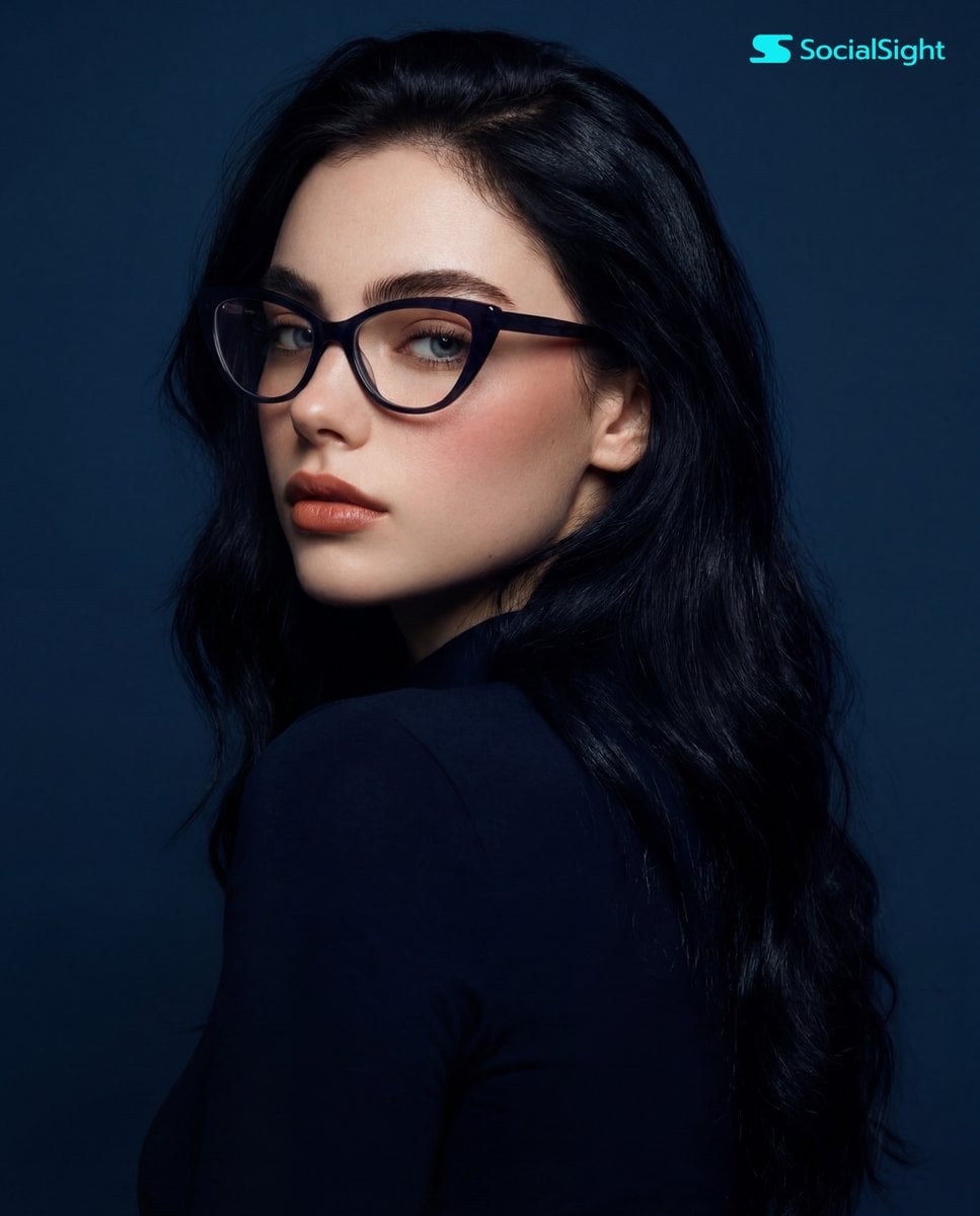 A young woman with long, dark hair and striking cat-eye glasses is captured in a studio portrait against a solid blue ba