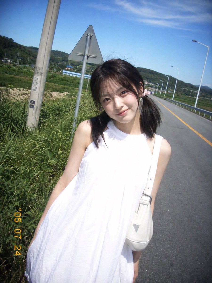 复古 CCD 卡片机夏日旅行抓拍，一位20岁出头的年轻东亚女idol站在乡间公路边。黑棕色低位双马尾，细碎刘海，浅粉色发圈，淡妆，冷白皮，真实皮肤。白色无袖高领宽松连衣裙，轻薄棉质，右肩白色单肩包。身体微微向右倾斜，头轻歪向左，直视镜头，自