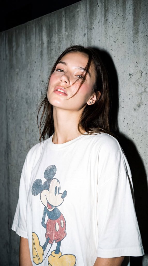 A high-resolution, close-up editorial-style portrait of a young woman standing against a textured gray concrete wall, ca