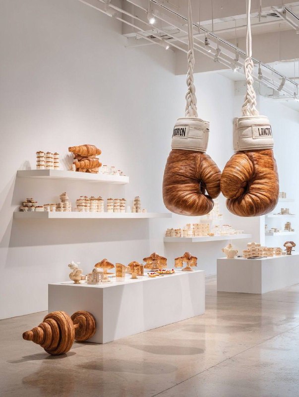 Minimalist white art gallery with surreal croissant sculptures. Center: oversize…